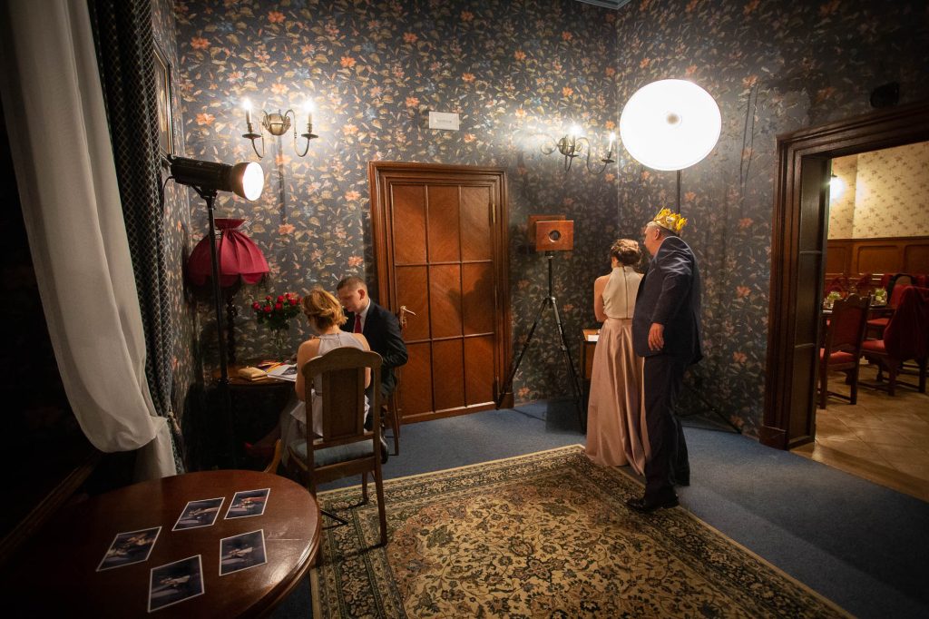 Fotobudka ustawiona w sali z tapetą w kwiaty: drewniany aparat w stylu retro na statywie, dwie lampy studyjne, goście i wydruki na okrągłym stoliku